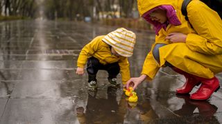 A little boy and his mother play in the rain on a paved walkway, both wearing yellow raincoats